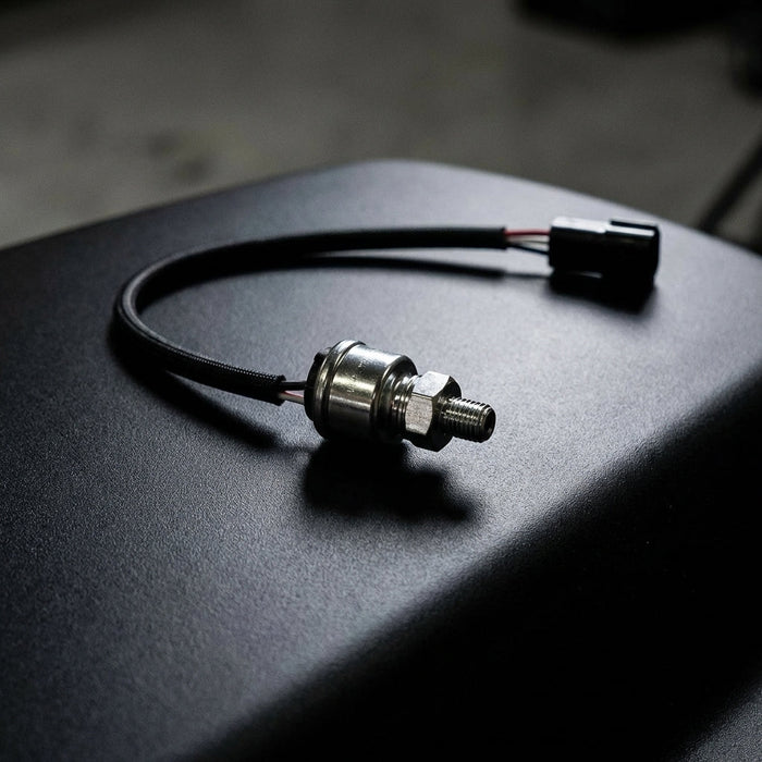 Full view of a small cylindrical fuel and oil pressure sensor with polished finish, placed on a sleek dark surface with dramatic lighting highlighting engineering details.
